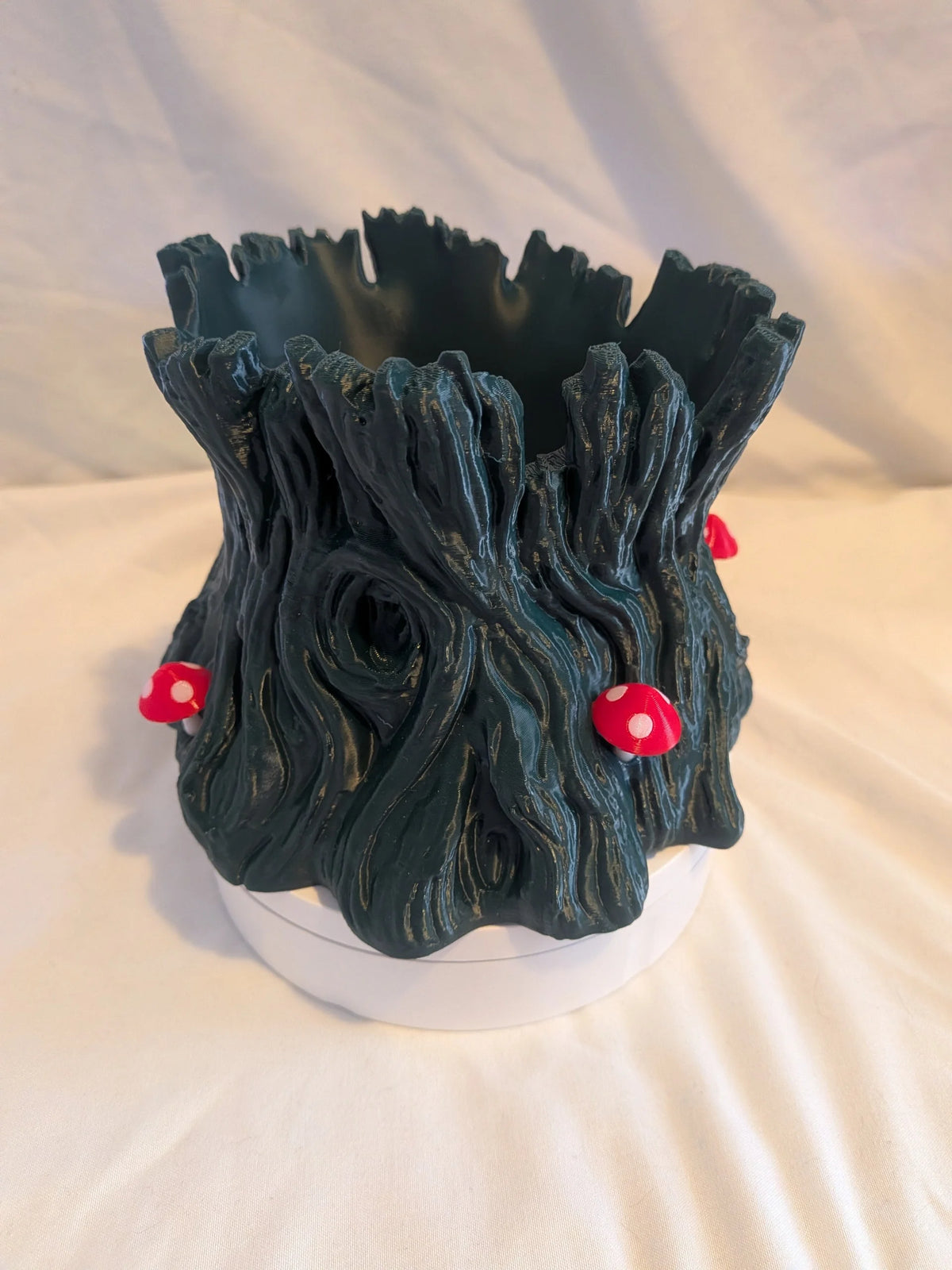 A dark green, tree-stump-like planter with a textured bark design and small red and white mushroom accents, displayed on a white circular base against a white fabric backdrop.