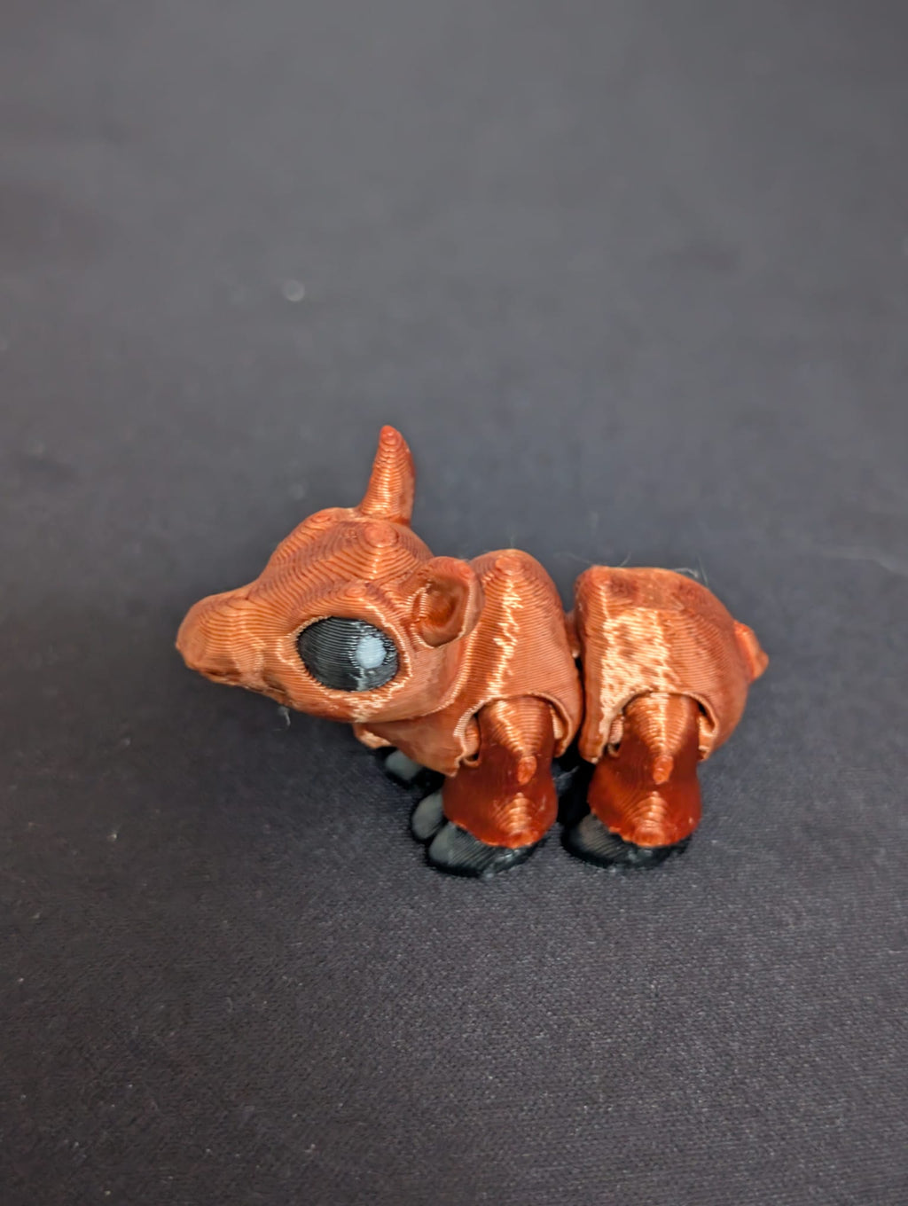A small Moose figurine with a metallic copper-brown body, black hooves, and a large glossy black eye, set against a dark textured background.