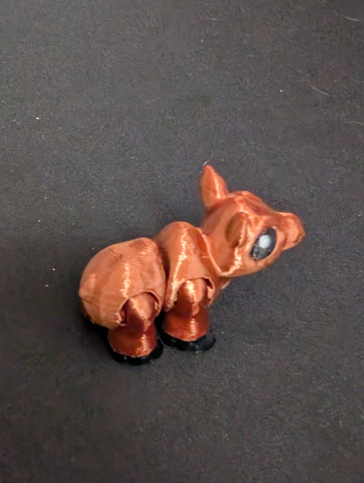 A small Moose figurine with a metallic copper-brown body, black hooves, and a large glossy black eye, set against a dark textured background.