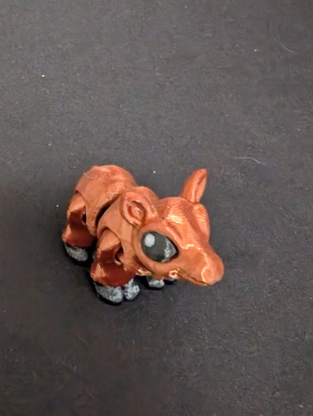 A small Moose figurine with a metallic copper-brown body, black hooves, and a large glossy black eye, set against a dark textured background.