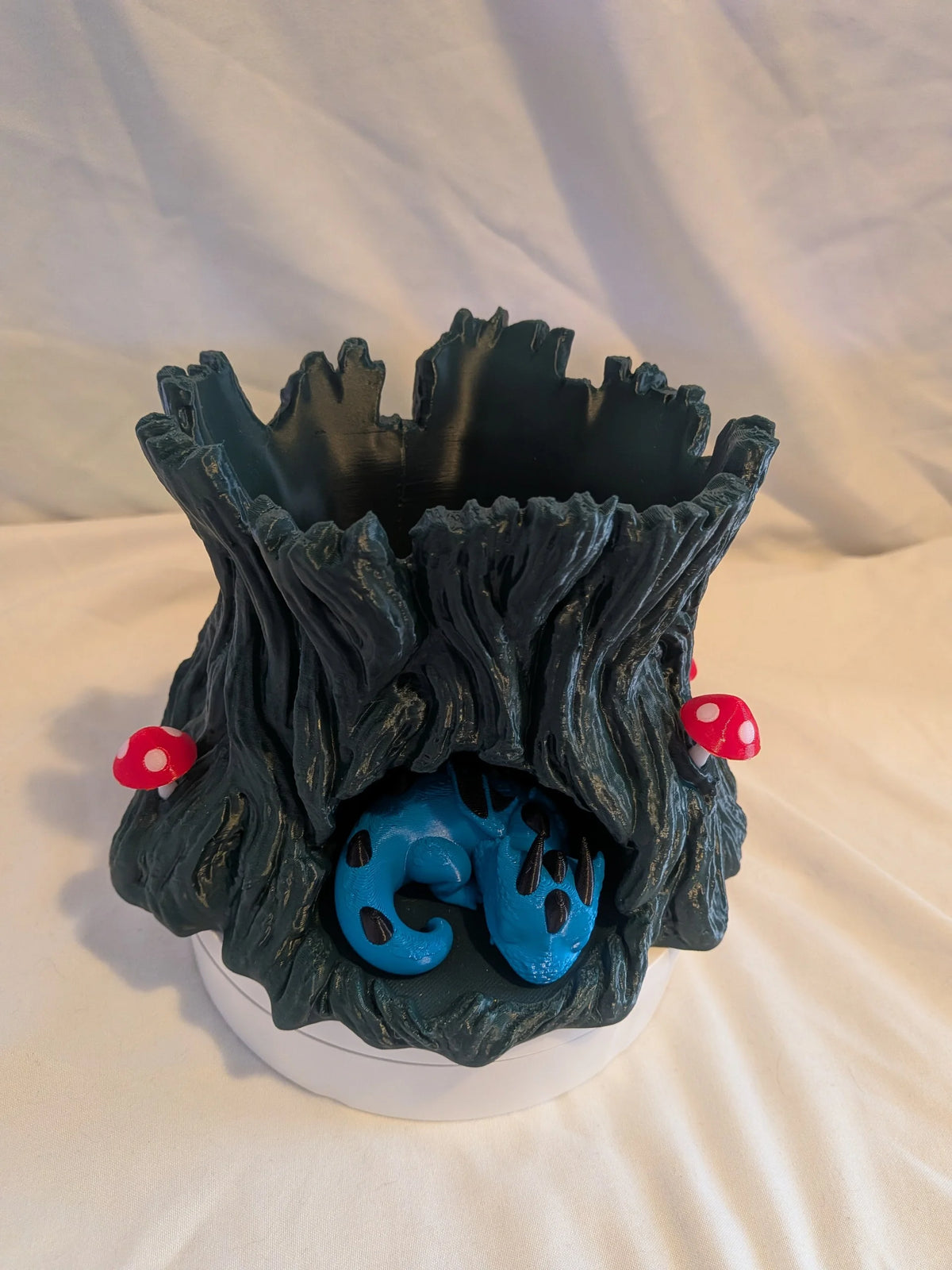 A decorative tree stump planter with a dark textured bark design, featuring a blue ladybug figure on the front and small red and white mushrooms on the sides, displayed on a white surface against a draped white fabric background.