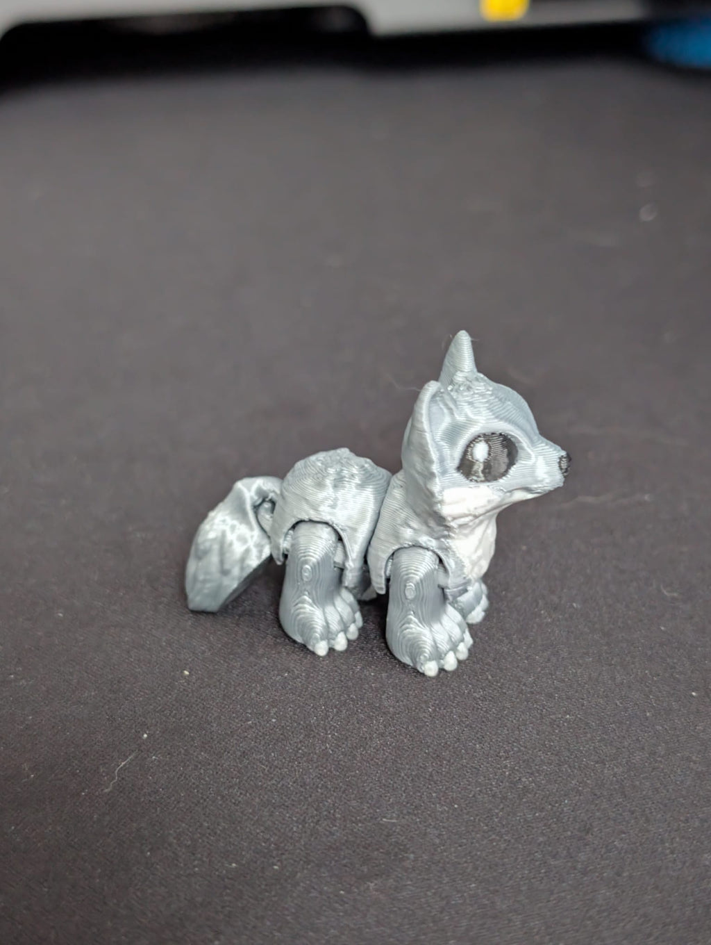 A small silver-gray articulated animal figurine, resembling a fox or chipmunk, sitting on a dark gray surface.